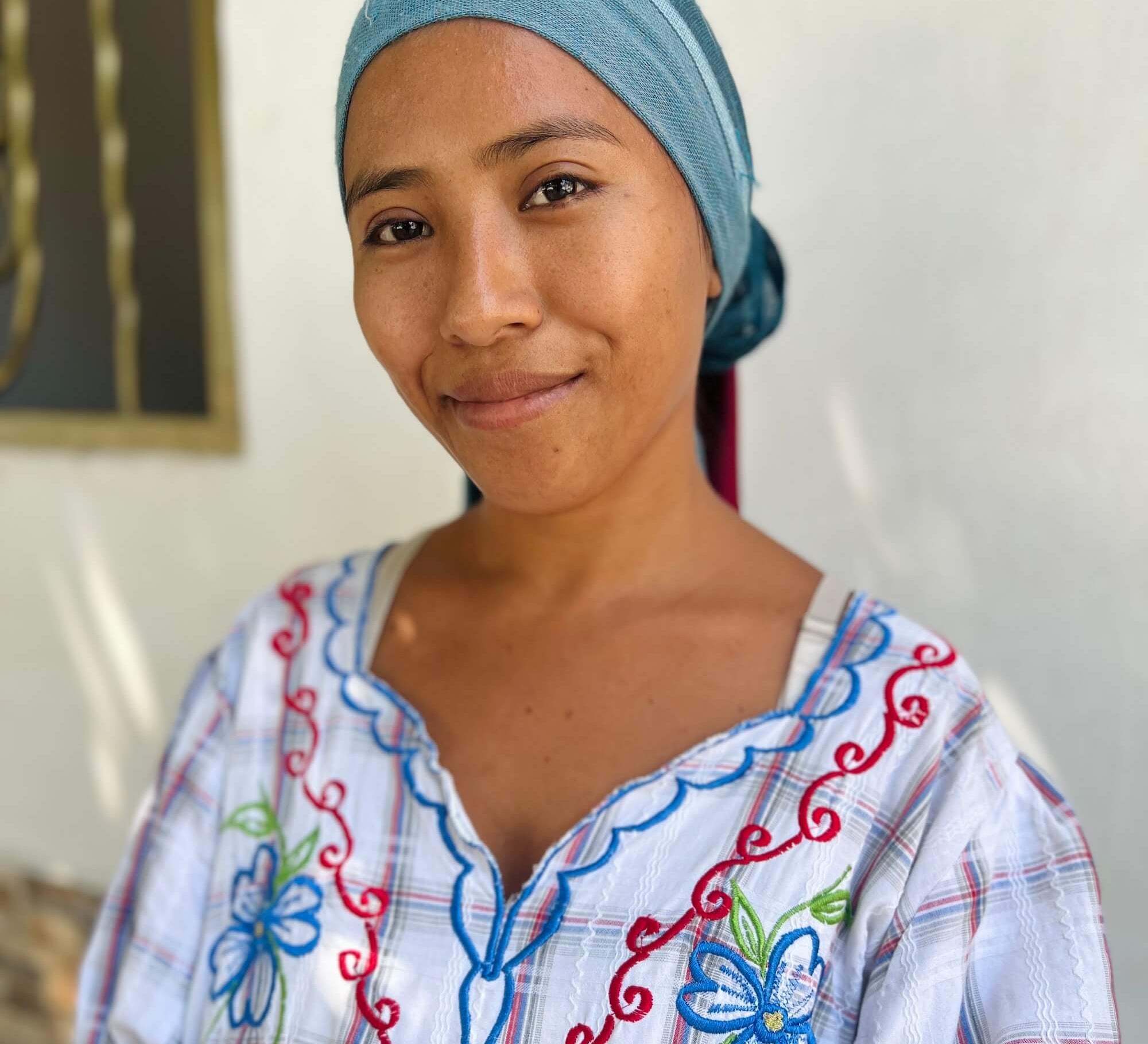 Woman wearing a colorful embroidered shirt and blue headscarf indoors.