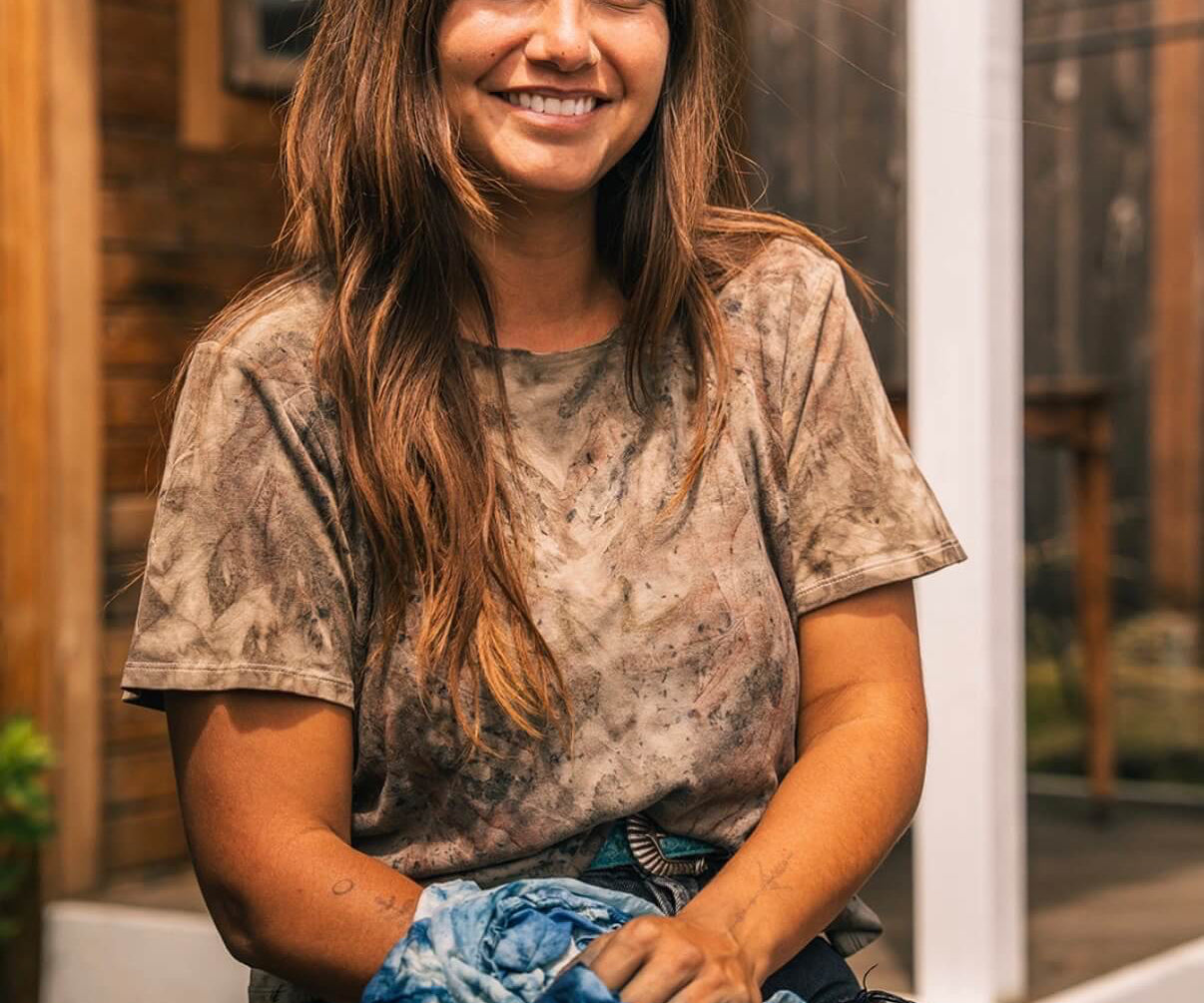 A smiling woman with long brown hair sits outdoors wearing a brown and beige tie-dye t-shirt and holding a blue and white tie-dye fabric bundle.
