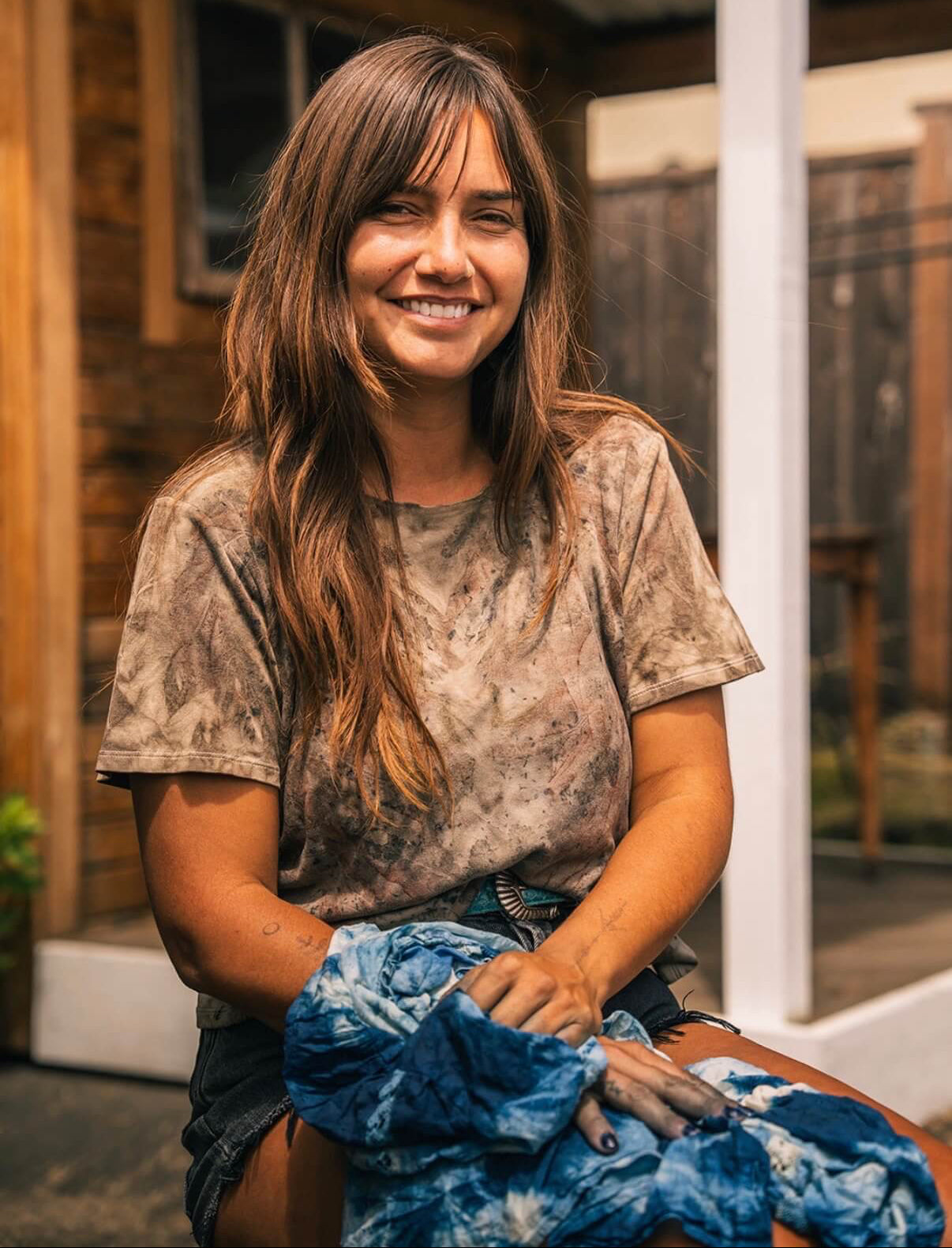 A smiling woman with long brown hair sits outdoors wearing a brown and beige tie-dye t-shirt and holding a blue and white tie-dye fabric bundle.

